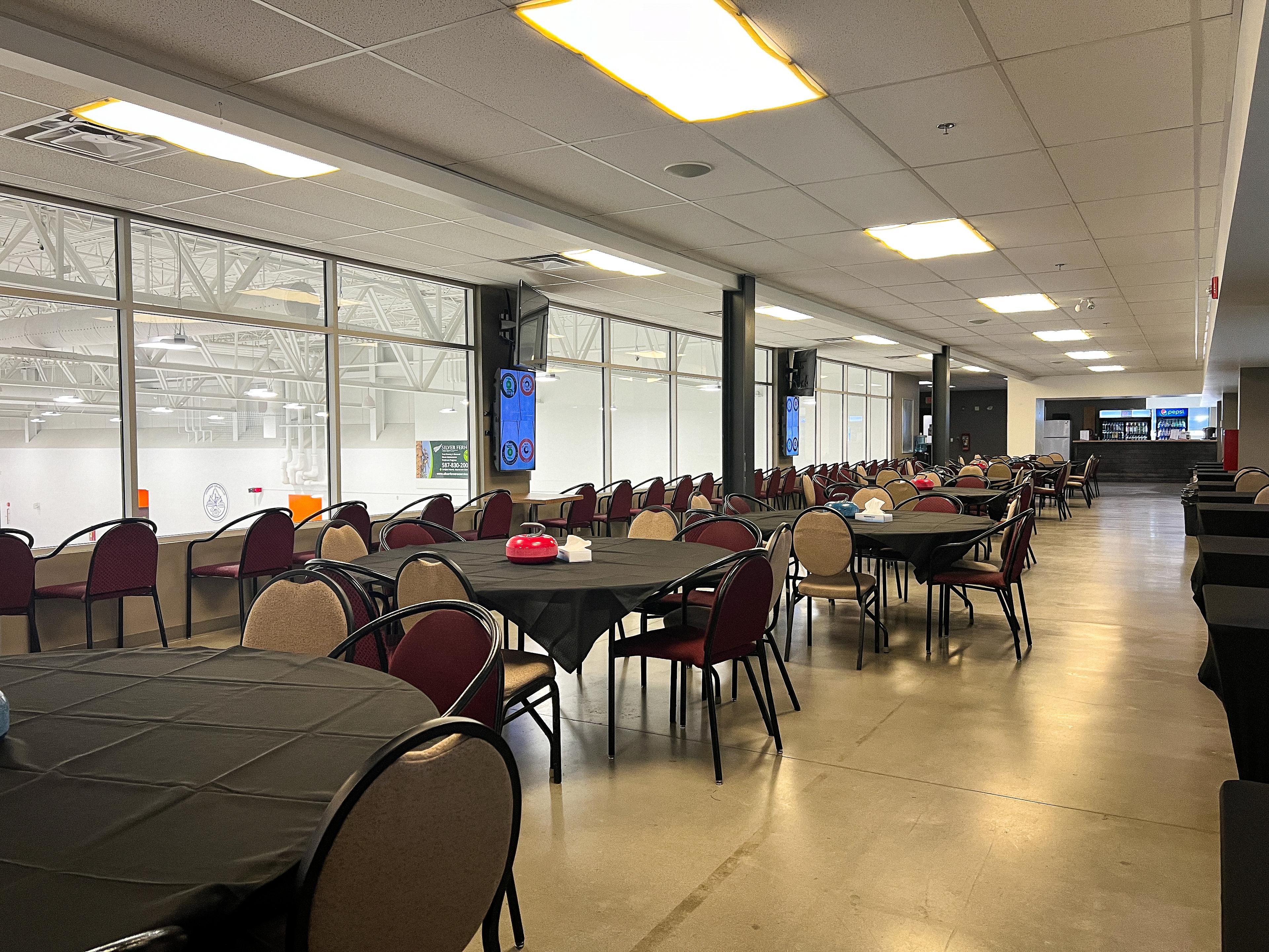 A lounge with round tables and chairs next to a wall of glass facing a curling rink. Every table has a curling rock and a box of tissues on it.