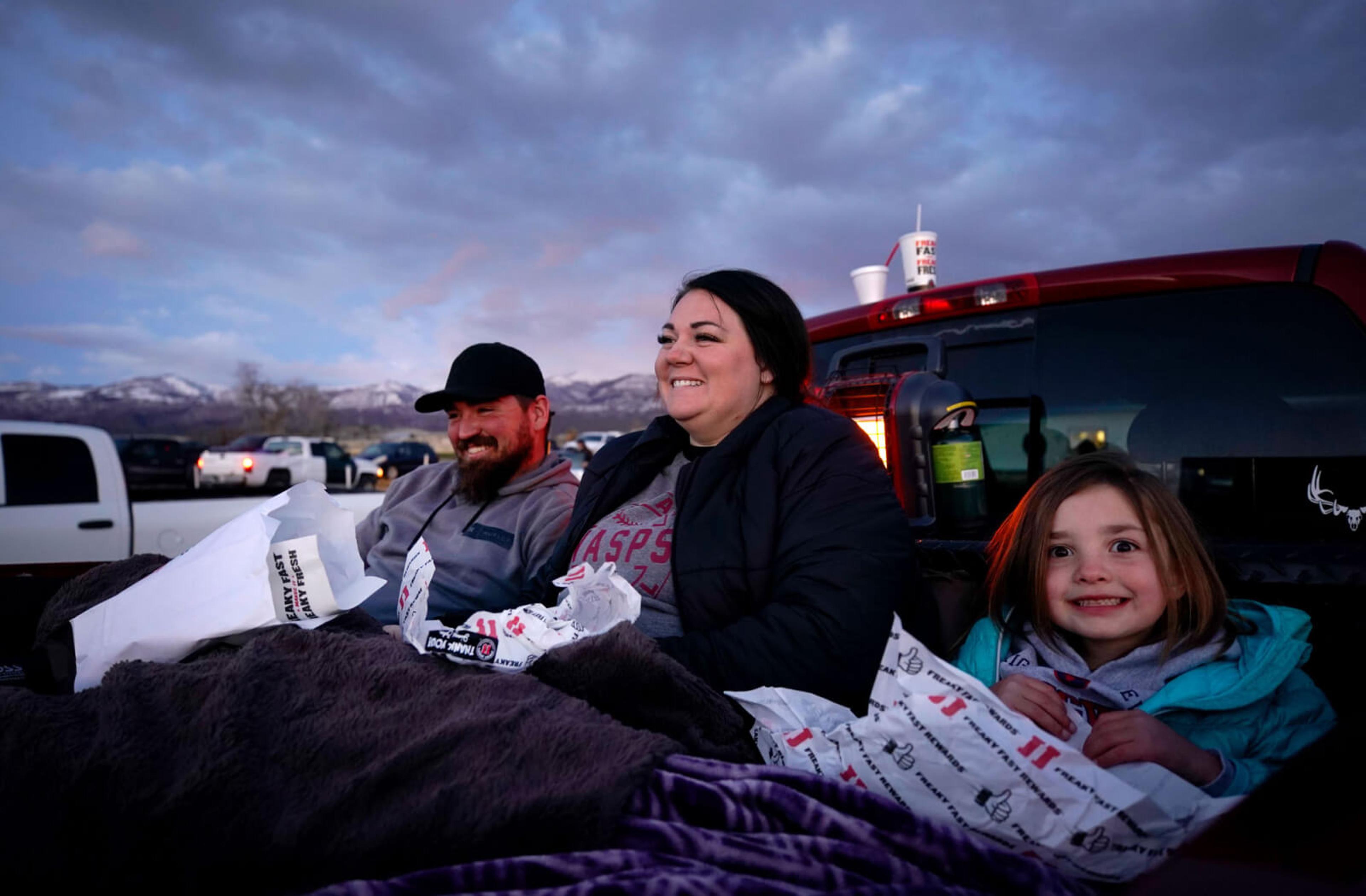 Three family members eat food in the back of their truck while attending an outdoor event.