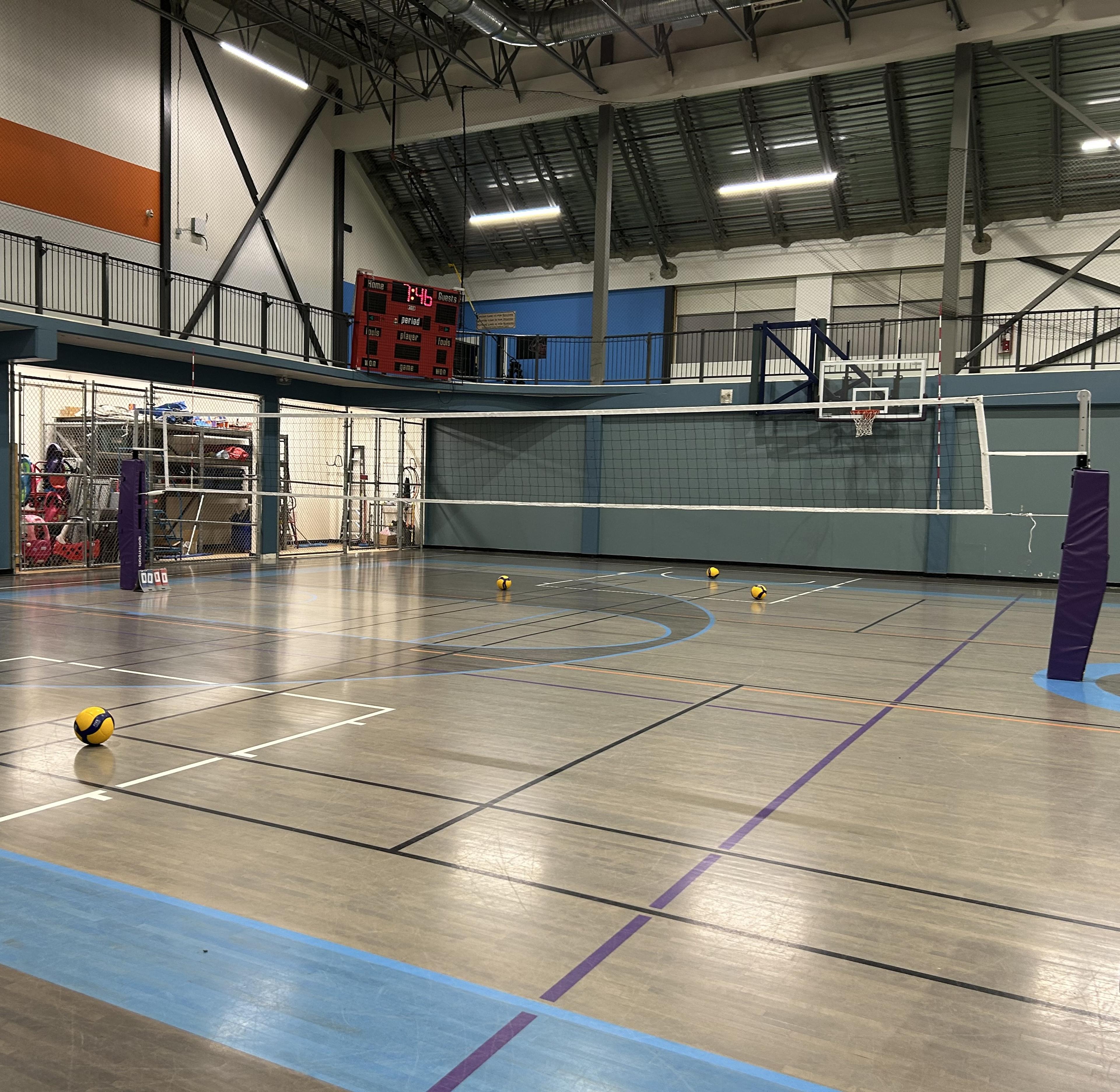 This is an image of a gymnasium showcasing a Volleyball net and some Volleyball balls scattered on the court.