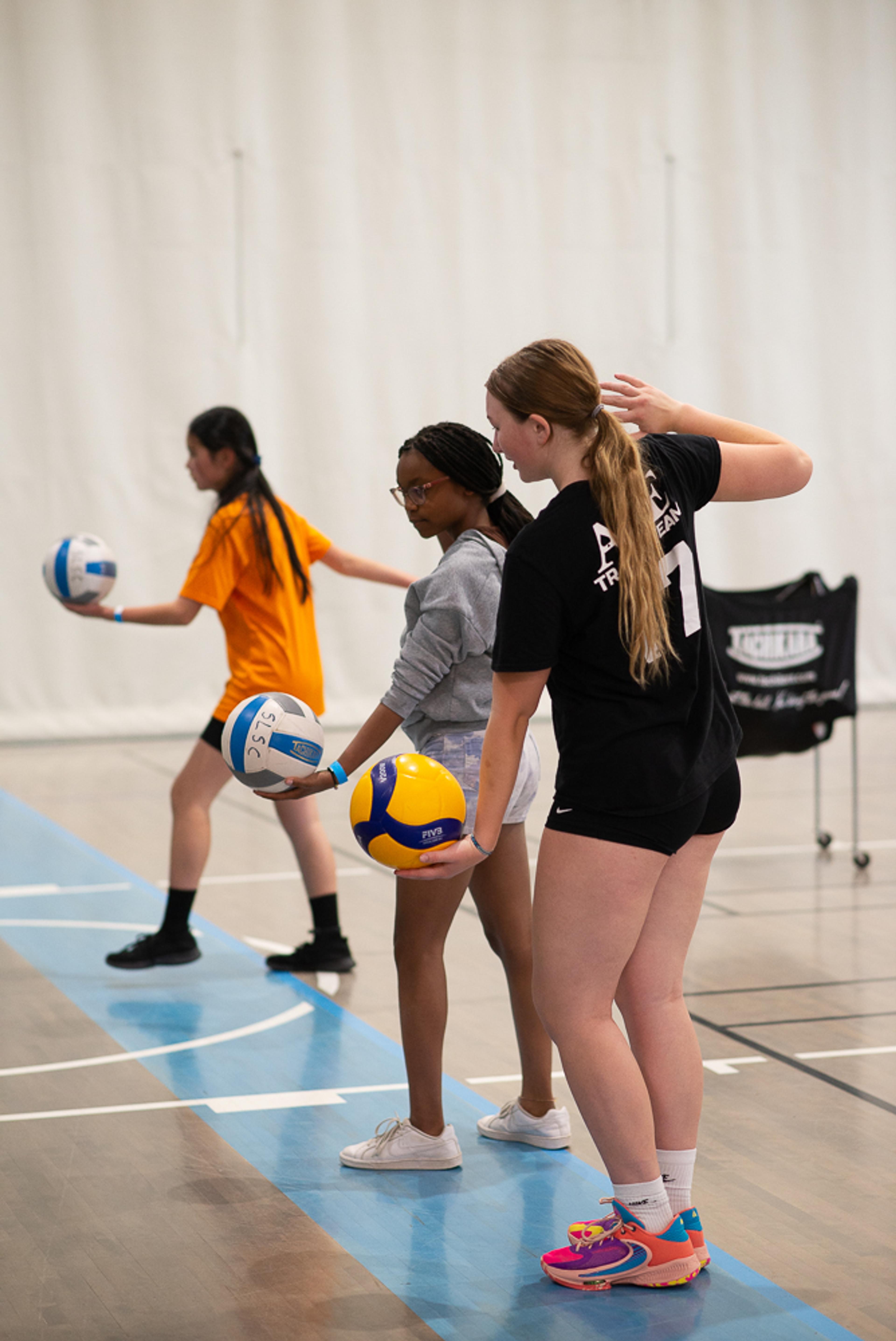 Young women being coached on serving a volleyball in a gymnasium.