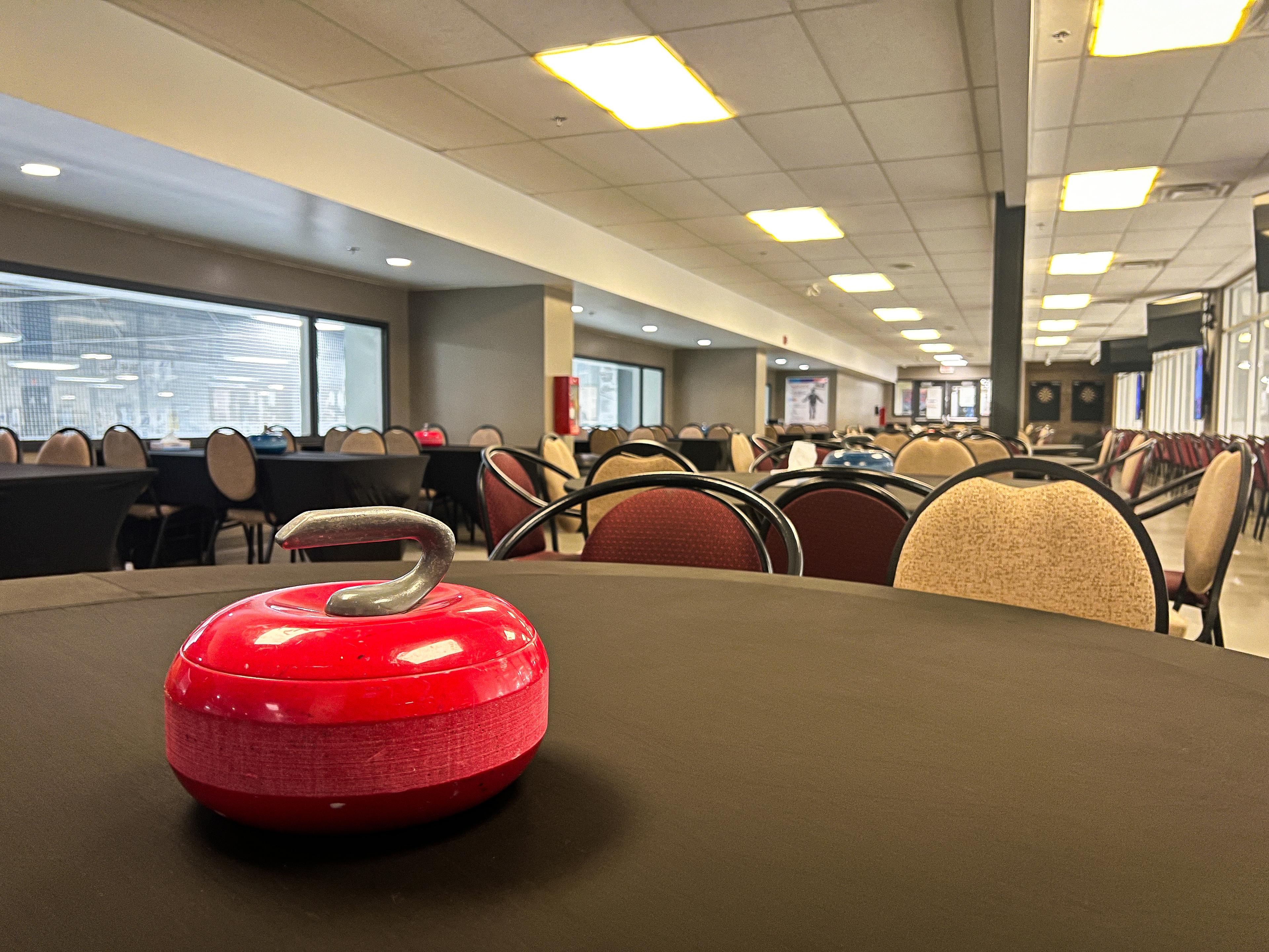 A close up of a curling rock on the table of a lounge with round tables and chairs next to a wall of glass facing a curling rink. Every other table has a curling rock and a box of tissues on it.