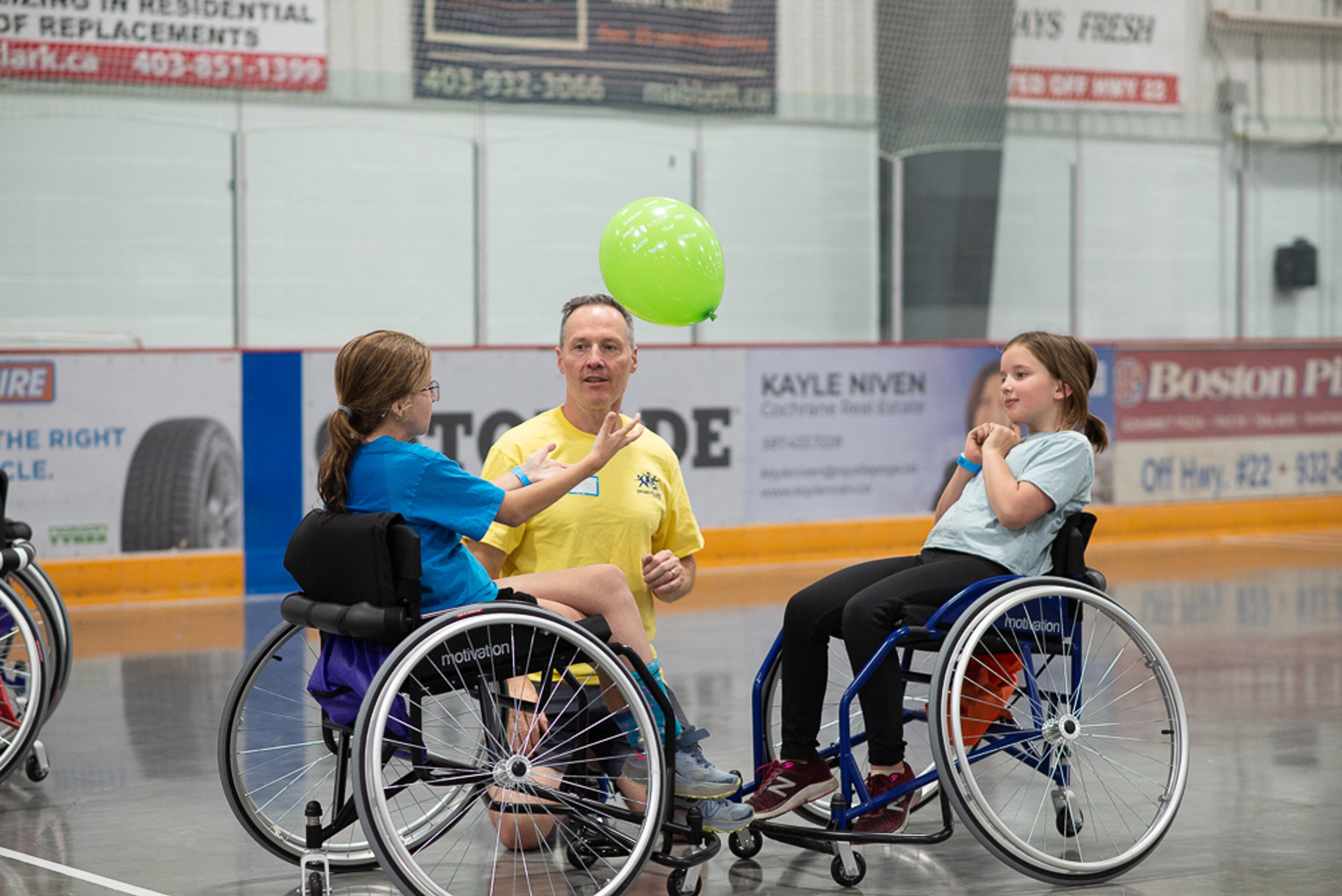 image of 2 young girls in wheelchairs playing with a balloon and a man kneeling helping