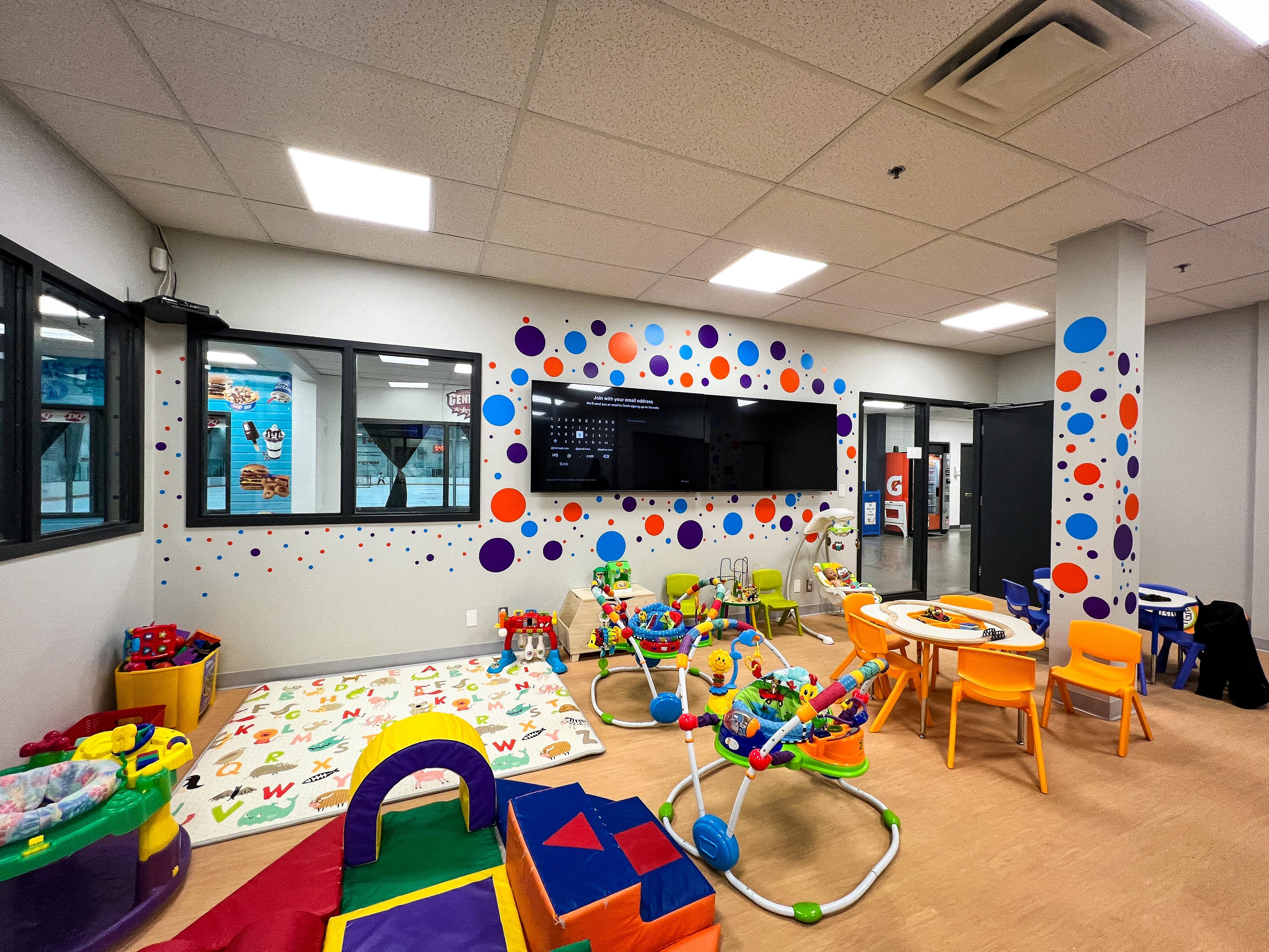 A Play care room with toys and kid size tables and chairs with 2 TVs and circle stickers on the wall.