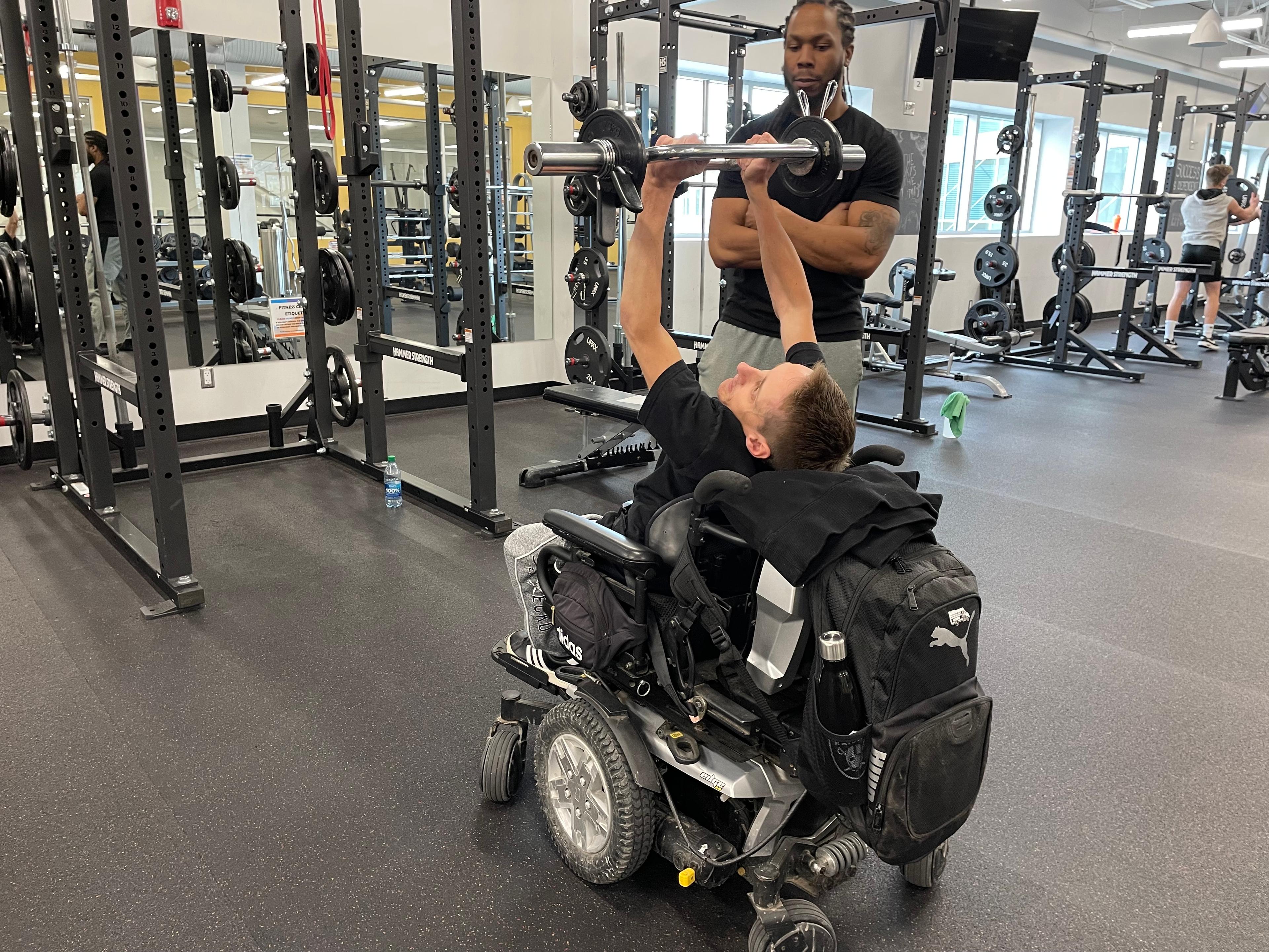 A picture of a man in a wheelchair lifting a barbell over his head, his personal trainer helping him out