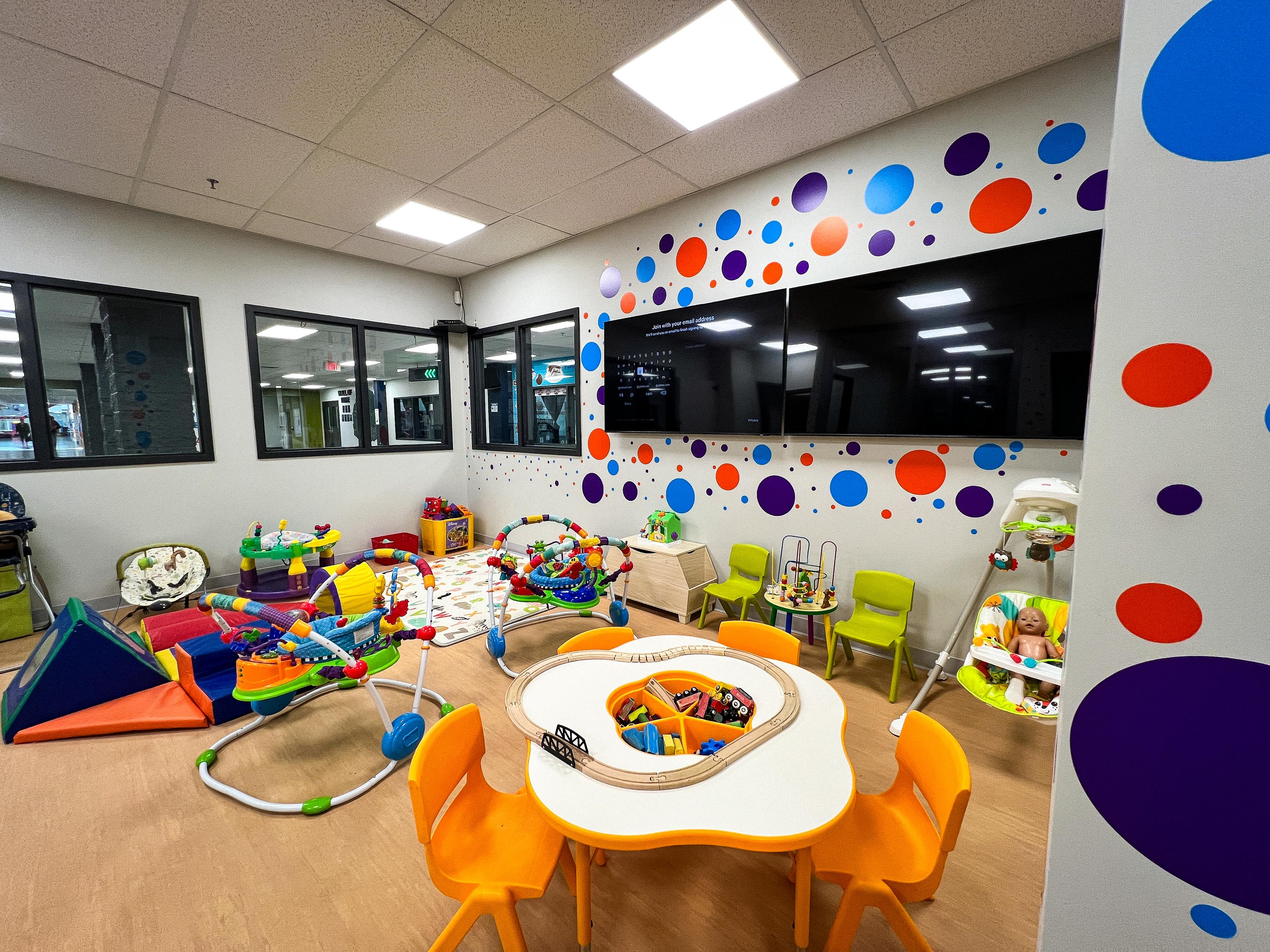 A corner of a Play care room with toys and kid size tables and chairs with 2 TVs and circle stickers on the wall.