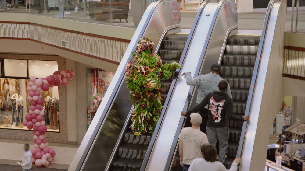 A man covered in vegetables descends a mall escalator and reaches to touch the hands of the people going up the escalator