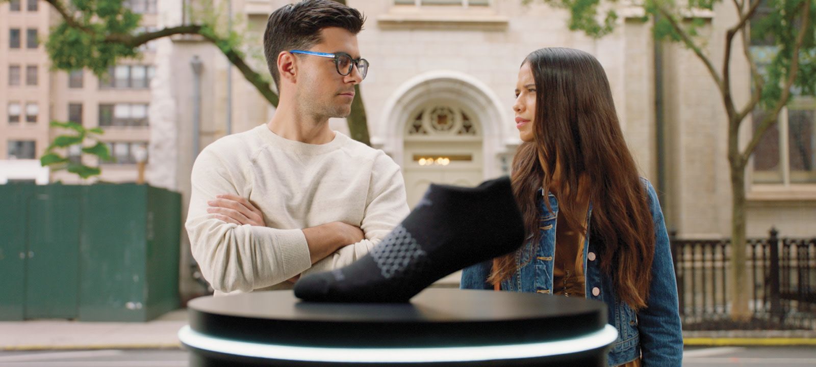 An older man and a younger girl look at each other puzzled from behind a sock on a pedestal close to the camera with a downtown city behind them