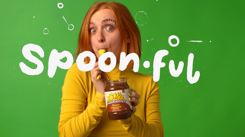 A woman with a yellow shirt in front of a green background eats a spoon full of sunflower butter out of the jar and the words, "spoonful" are written across the screen in white