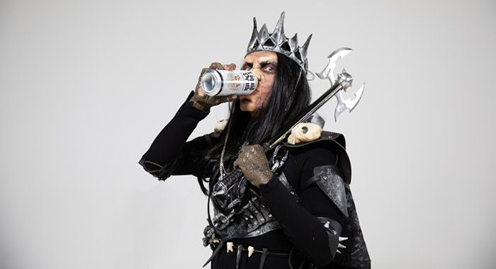 A man in an all black monster costume with paint and a crown drinks liquid death and wields an axe