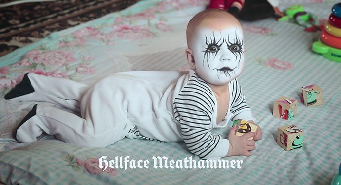 A baby laying on its stomach on a blanket on the ground surrounded by toys looks up with black and white facepaint and the caption under him says, "Hellface Meathammer"