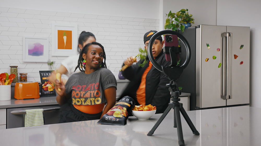 a man and woman eat a bowl of terra chips in a kitchen as a phone records them on a tripod on the counter