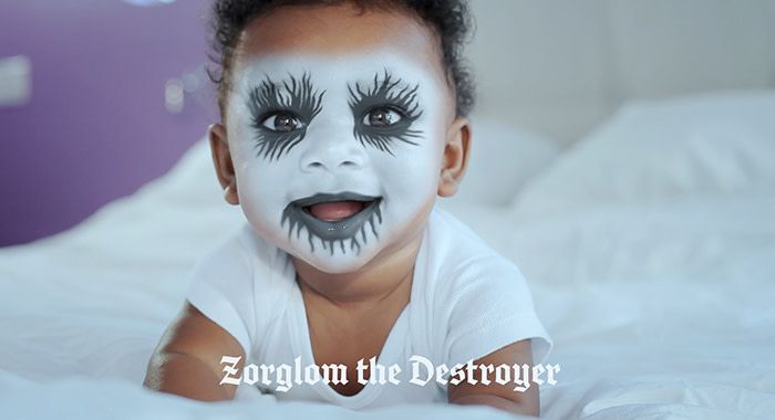 a baby in bed with black and white face paint looks into the camera and the caption under his face reads, "Zorglom the Destroyer"