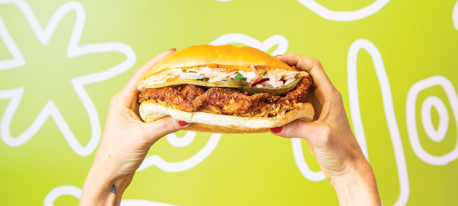 A downward shot of hands holding a holy chicken sandwich with a green back ground with wavy symbols