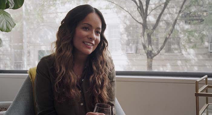 A woman sits and looks just past to the right and holds a wine glass smiling and the window and a tree is behind her