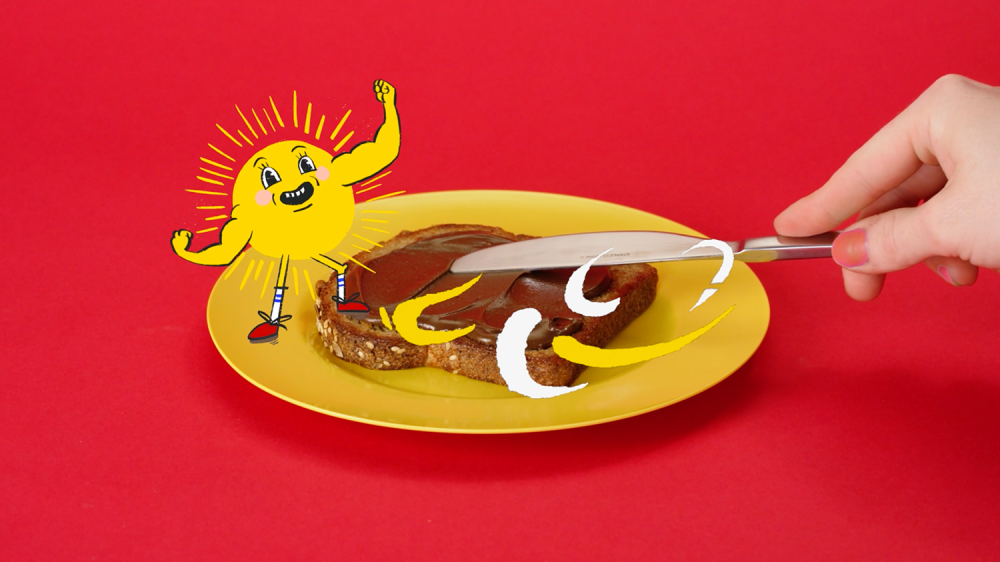 A close up shot of a hand spreading sunbutter on toast with a knife is shown as a graphic of a sun flexing his muscles is displayed over top of the image