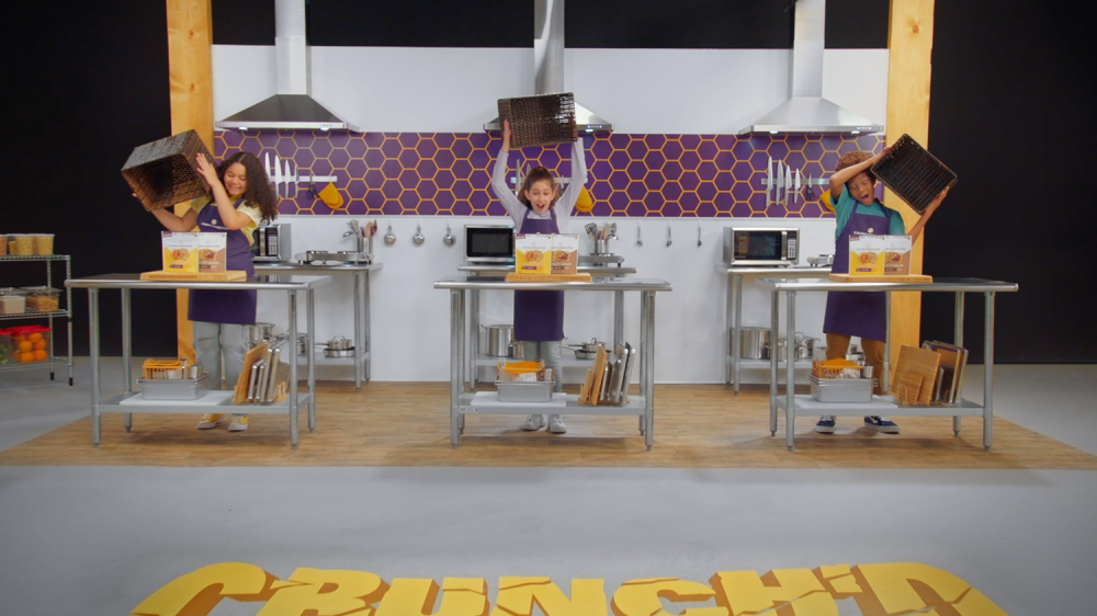 3 children in a kitchen set take boxes off of the table to reveal boxes of crunchmaster crackers