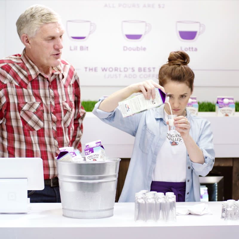 A woman pours our half and half into a glass in a coffee shop with directions behind her and another man watches her from the side