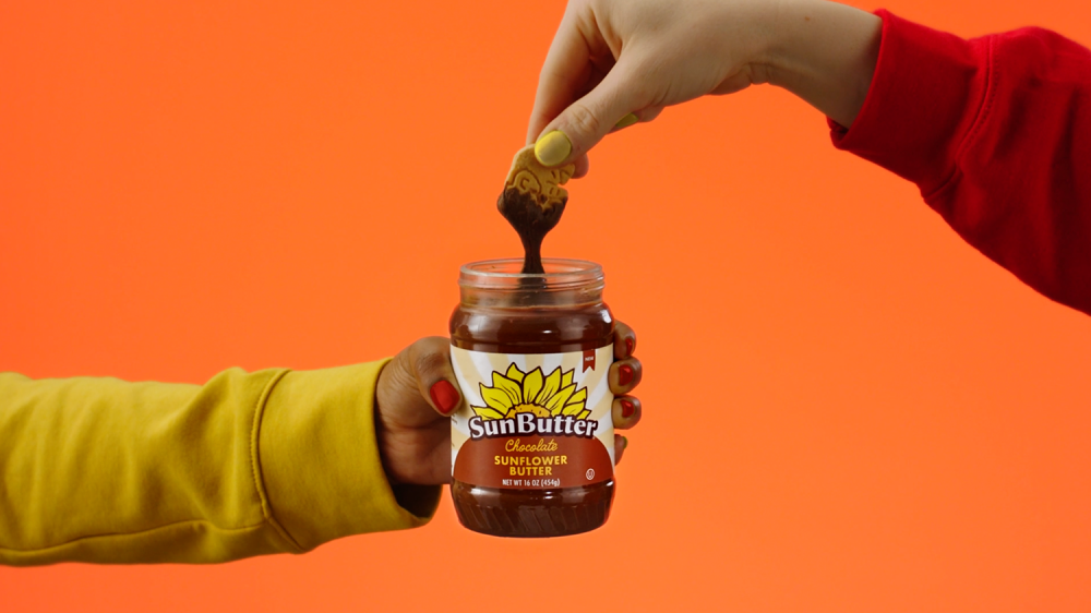 One hand from the left holds the sunbutter jar while a hand from the right dips an animal cracker into the sunbutter jar