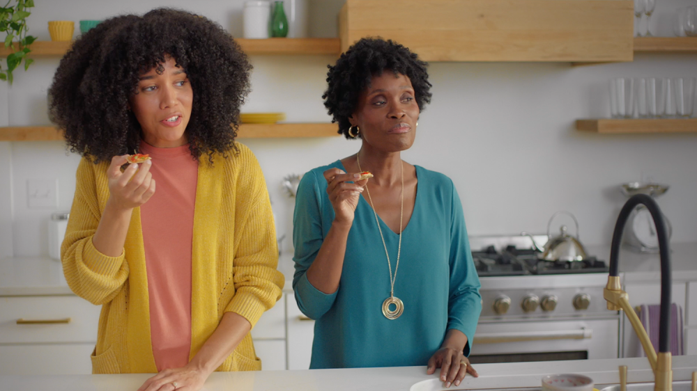 Two women in a kitchen eat crackers with toppings