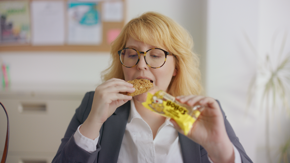 Woman in glasses takes a bite of a bobo bar in an office setting