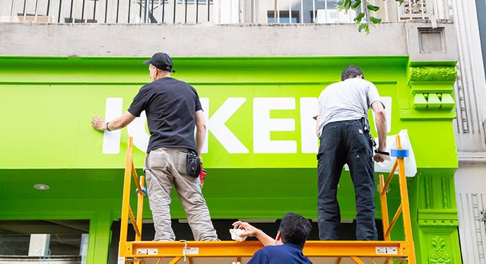 two men stand on scaffolding pasting letters on the top of a lime green store