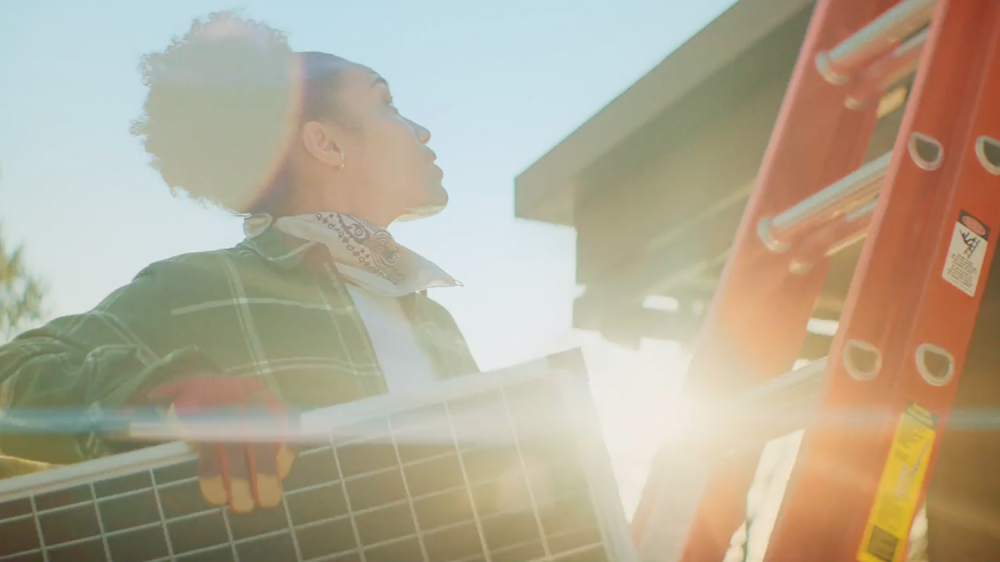 A close up shot of a woman about to climb a ladder holding a solar panel with the sun beaming through