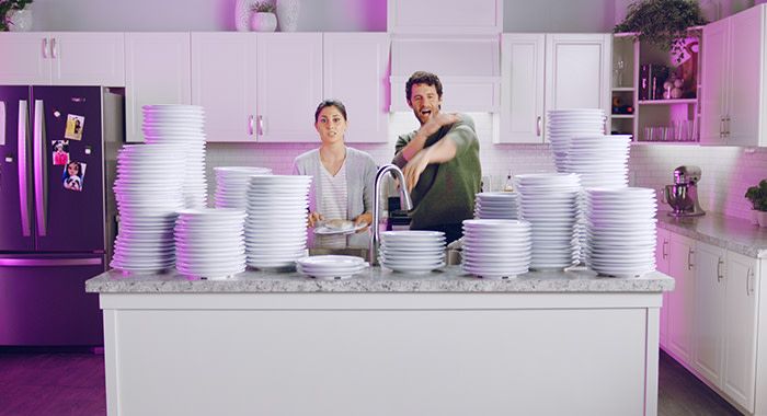 A man and a woman in a kitchen are surrounded by plates and there is a purple light in the background