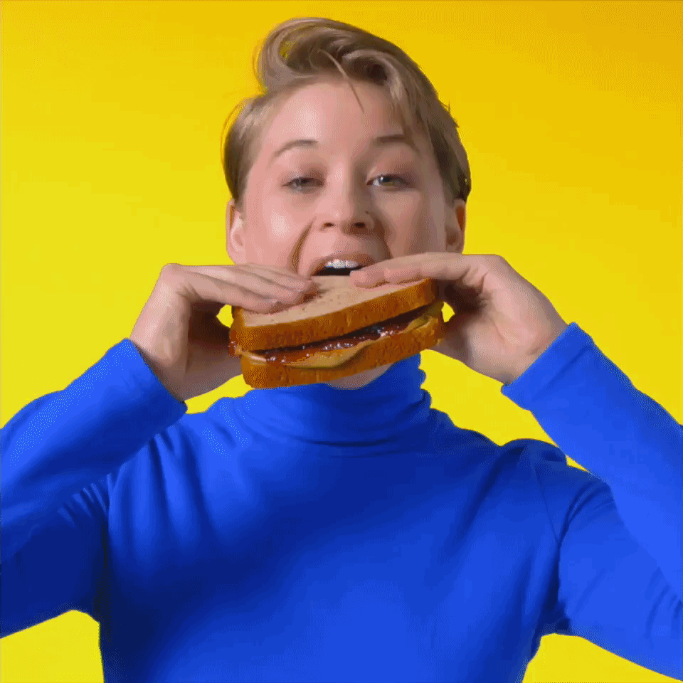 A gif of Multiple people each in brightly colored clothes and with a brightly colored background bite into a sunbutter and jelly sandwich with the words flying across, "Mmmmmmmmmmm"