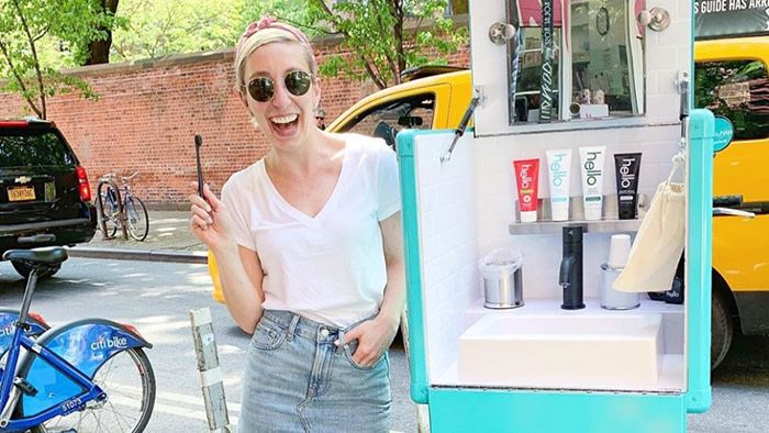 a woman holds up a toothbrush smiling next to the mobile hello teeth brushing station