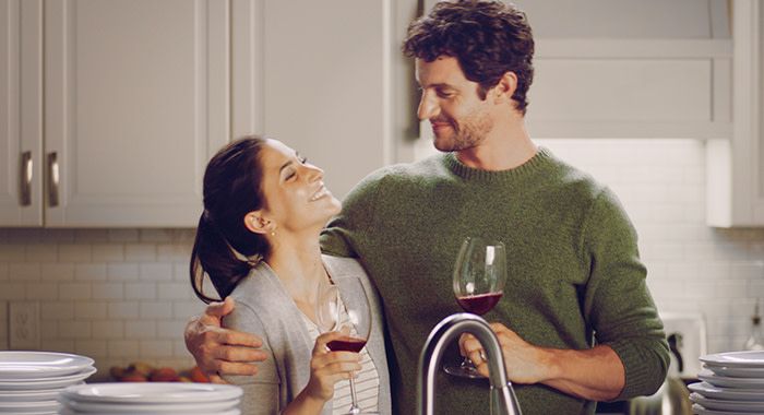 A man and a woman in a kitchen hold wine and the man puts his arm around the woman and they both smile