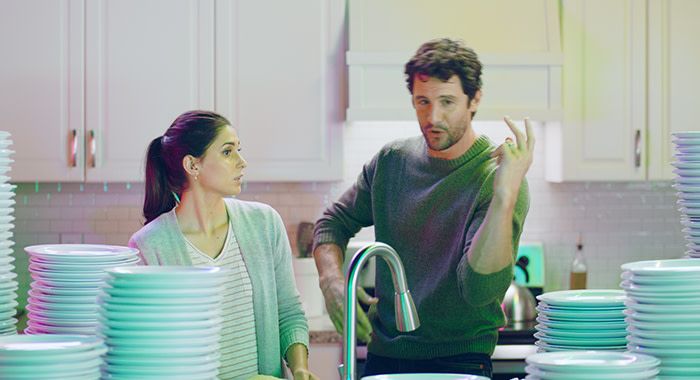 A man and a woman in a kitchen behind a sink are surrounded by plates and talk to each other