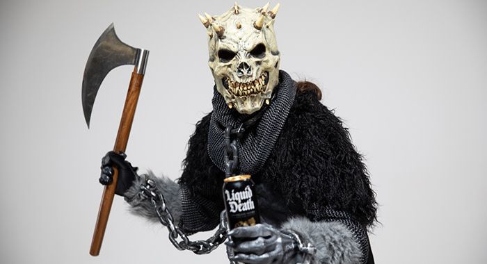 Man in Spiky headed monster costume holds liquid death in his left hand and an axe in the other while chained up