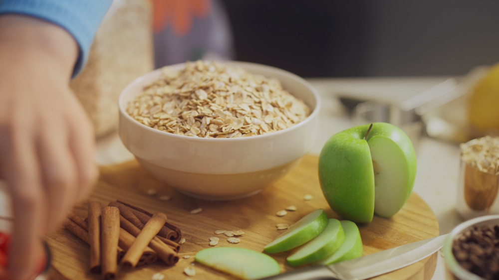 close up shot of an apple, cinnamon, and bowl of oats while a woman grabs other ingredients to mix together