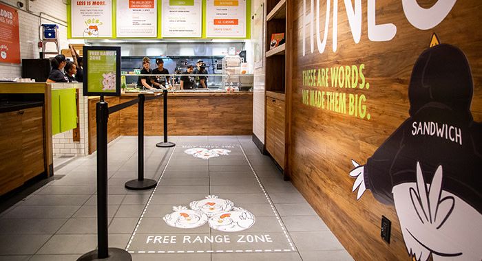 A wait line for the Holy Chicken restaurant with a drawing on the ground where people wait in line that has chickens and reads, " free range zone