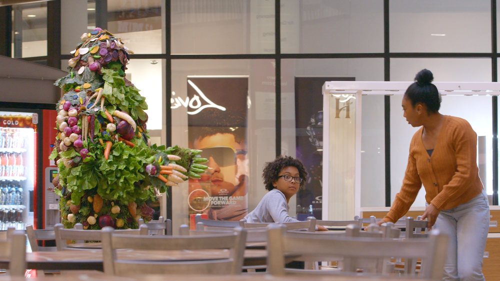 A man covered in vegetables walks around the food court approaching a woman and child