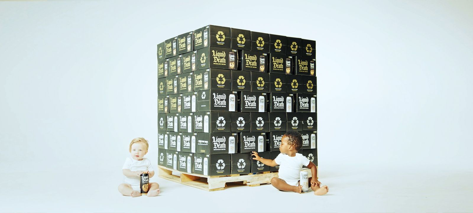 Two babies sit in front of a pallet holding many boxes of liquid death mountain water and the baby on the left holds a can of liquid death