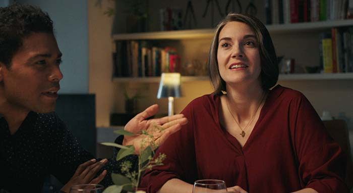 woman sits next to a man at dinner table with bookshelf and lamp behind her looking up and to the left