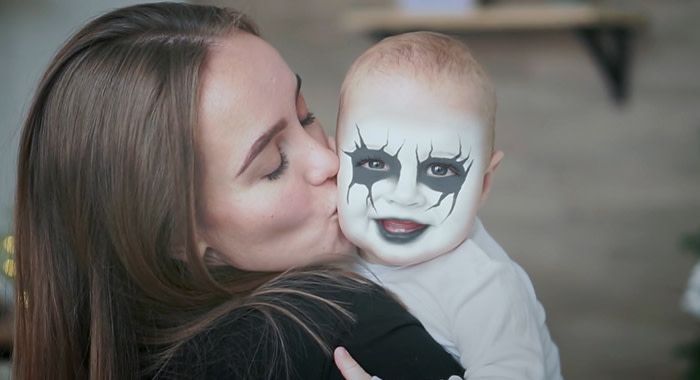 A woman is holding and kissing a baby with black and white facepaint around its eyes and mouth