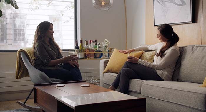 two women sit on a couch drinking wine looking at each other talking