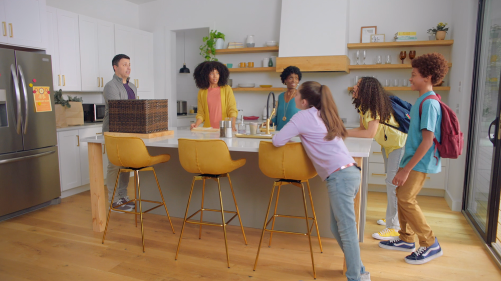 Children with backpacks on coming into a kitchen talking to adults around a kitchen counter