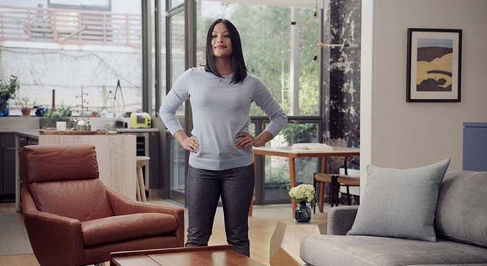 a woman stands in her living room next to her couch with the windows behind her, with her hands on her hips and a smile on her face