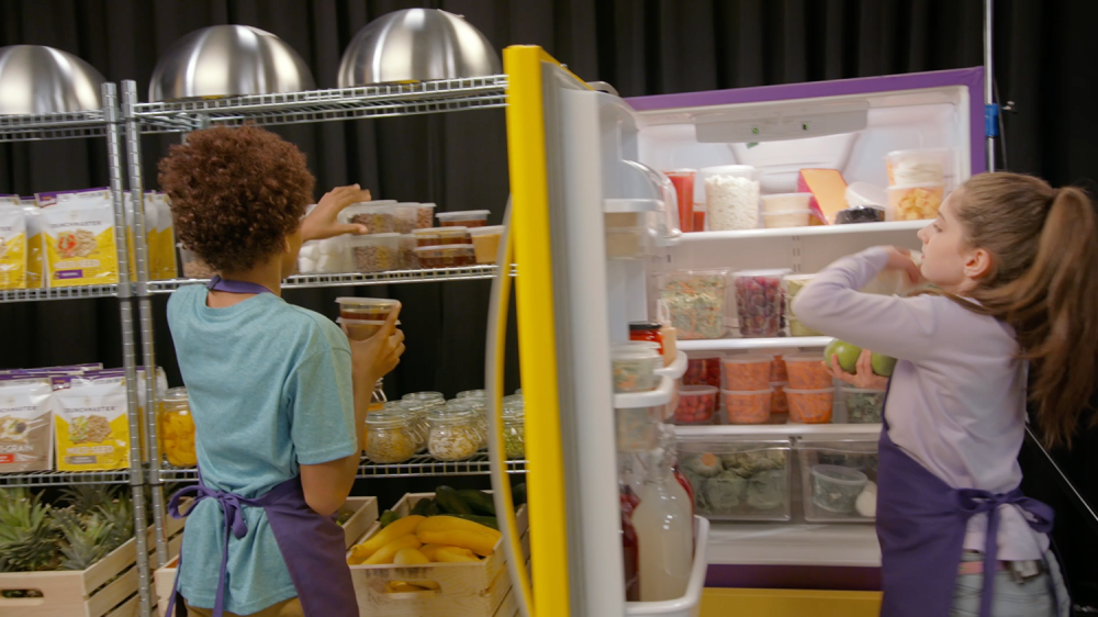 2 Children with aprons grab containers of food out of refrigerator and shelf