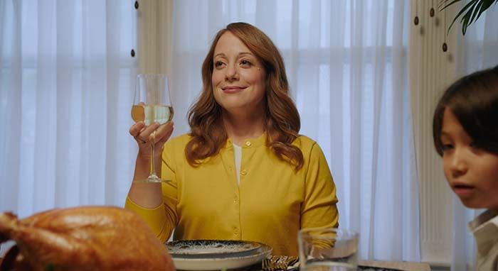 woman sits at table with curtains behind her and turkey in front of her raising her wine glass and smiling