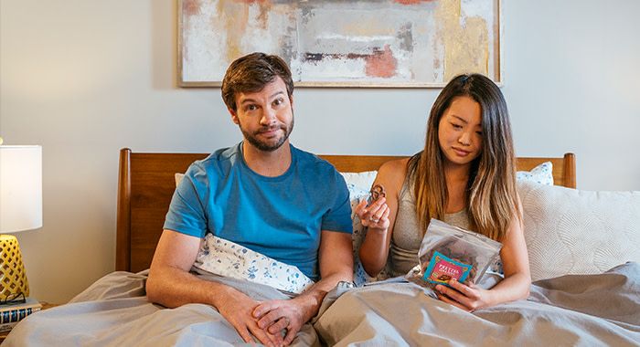 A man and woman sit up in bed while a man looks at the viewer with a smug look while the woman looks confused at a bag of pretzels