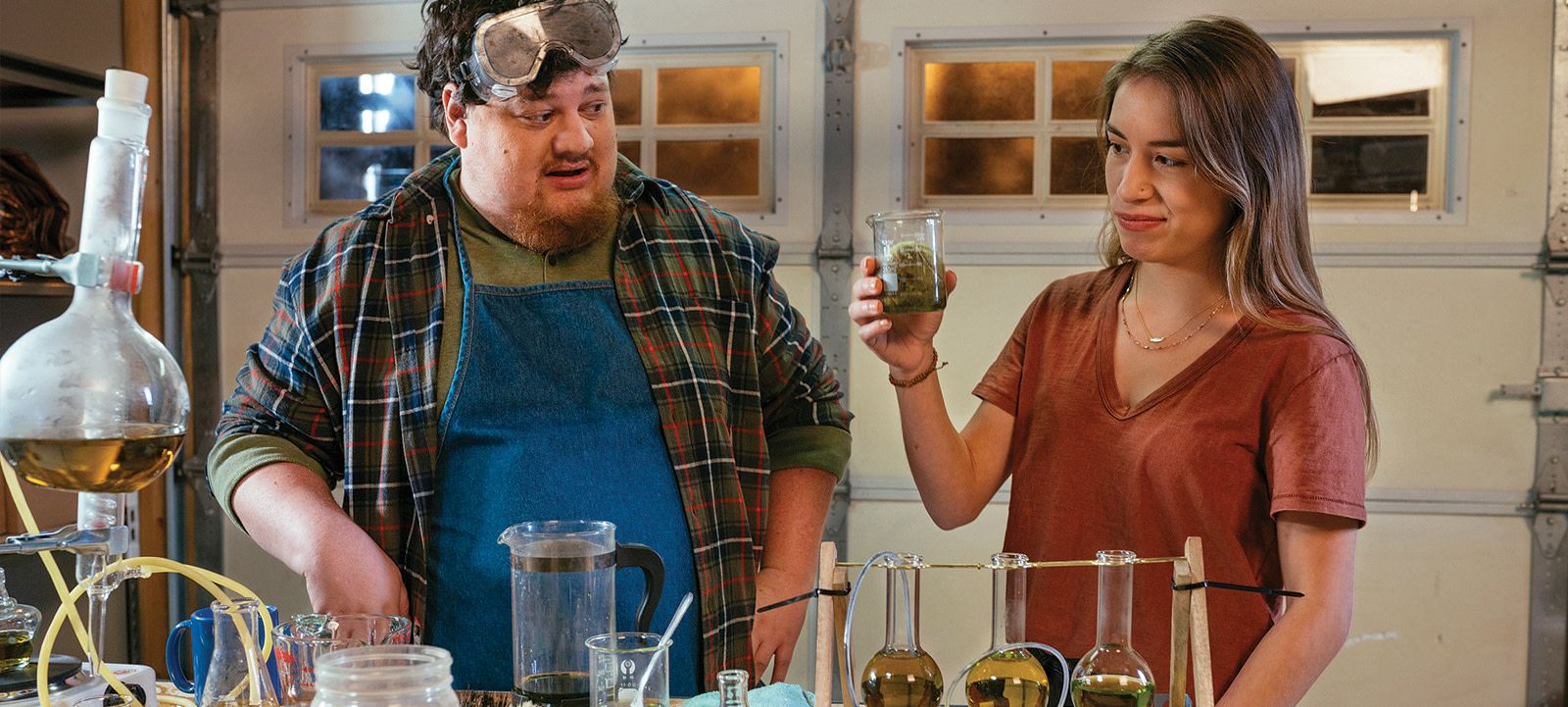 a man and woman look at a soggy tennis ball in a beaker surrounded by a garage chemisty set