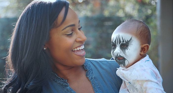 A woman is smiling while holding her baby outside and the baby has black and white facepaint