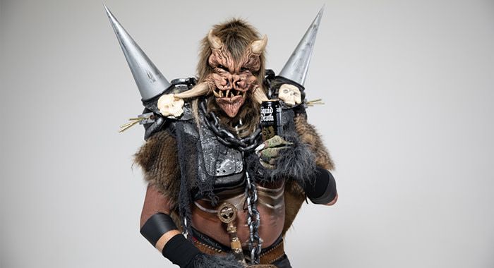 Man in monster costume with spikes coming out of his face and shoulders holds a black can of liquid death