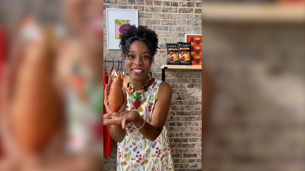 A woman holding a sweet potato close to the camera in front of a brick wall