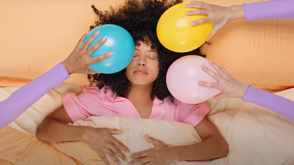 A woman with curly hair laying in bed and multiple hands come from outside the picture holding balloons rubbing it on the sleeping girl's hair