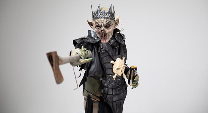 A person in a monster costume and crown holds a liquid death and points an axe at the viewer